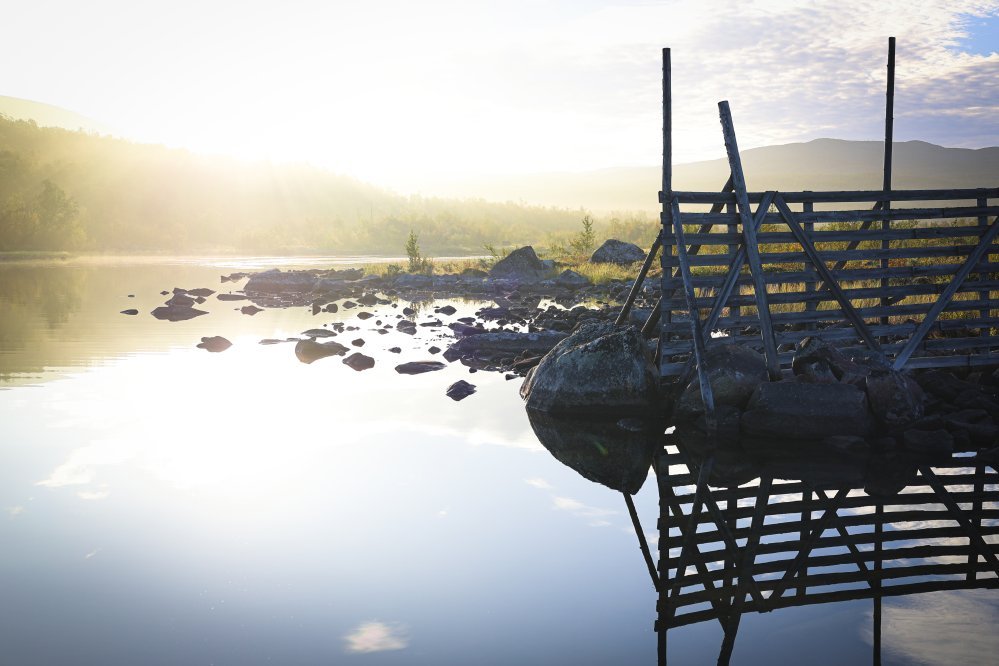 Kuvassa on vanhaa tuuliaitaa ja Kilpisjärven rantaa, taustalla tunturia sumussa ja juuri nousevaa aurinkoa