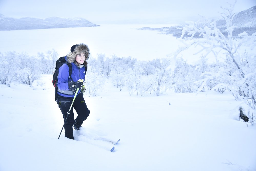 Nainen hiihtää Kilpisijärvellä lumisessa metsässä. Naisella on karvalakki päässään ja lila hiihtotakki. 