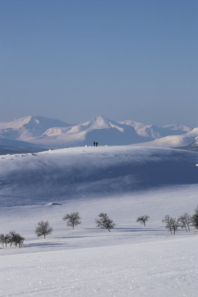 Kaksi hiihtäjää seisoo kaukaisuudessa lumisen maiseman ja Kilpisjärven suurtuntureiden keskellä