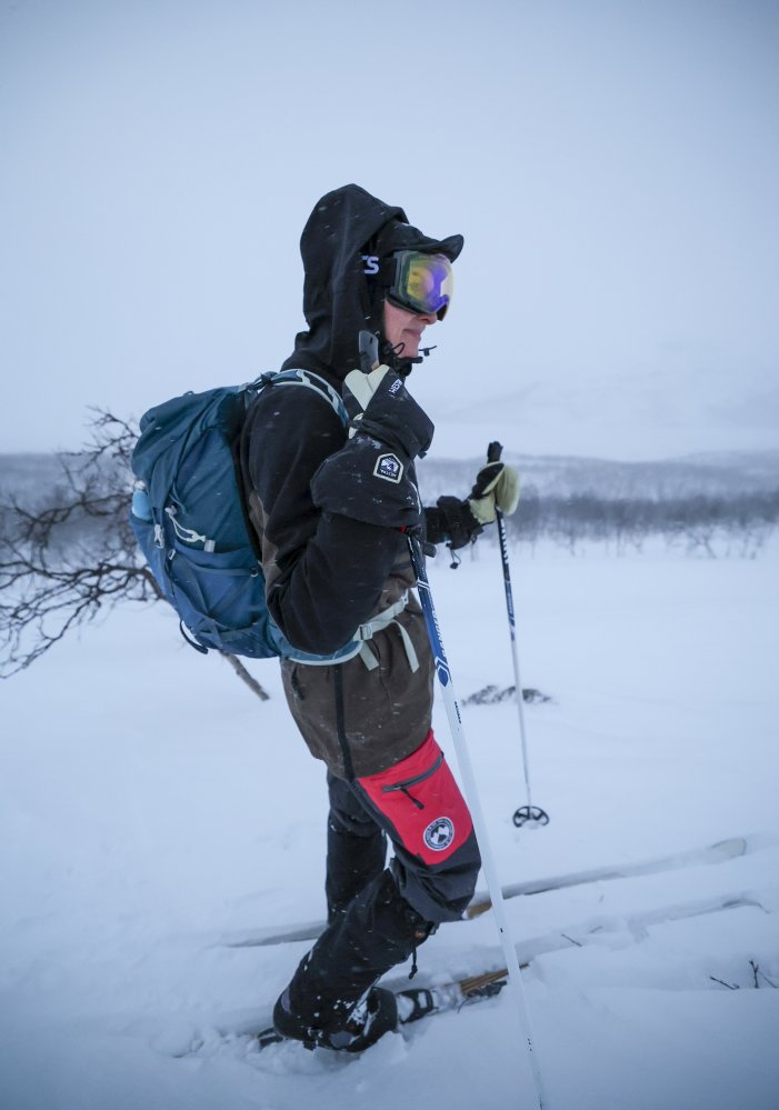 Nainen seisoo tunturin rinteellä kovassa lumituiskussa. Taustalla erottuu juuri ja juuri Pikku-Mallan tunturi. Nainen on suksilla ja kädessään hänellä on sauvat. Naisella on reppu, tuulelta suojaavat hiihtolasit, säärystimet jalassa sekä huppu päässä.