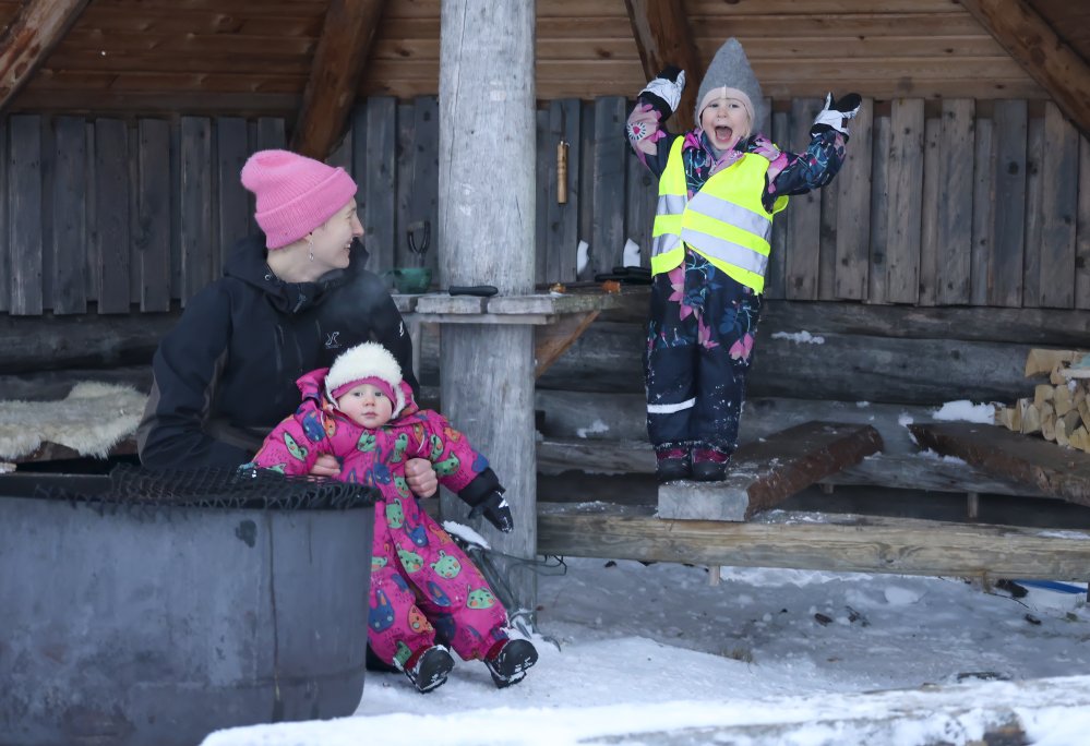 Lapsi seisoo talvihaalareissaan laavussa tulien äärellä. Lapsi huutaa ilosta ja molemmat kädet ovat nostettuina kohti kattoa