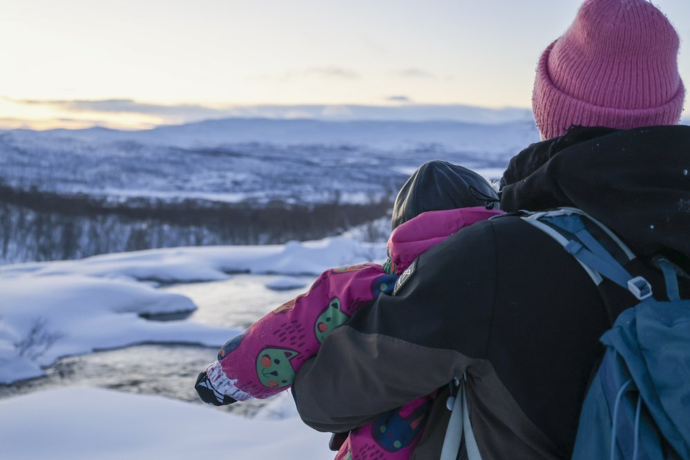 Kuvassa nainen sylissään lapsi katsoo edessään olevaa sulaa vettä ja puroa talvisena päivänä Tsahkaljärvellä