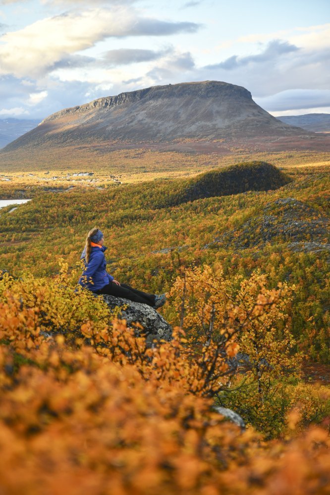 Nainen istuu tunturin rinteellä ja katselee värikästä ruskamaisemaa kohti Kilpisjärven kylää ja Saanatunturia