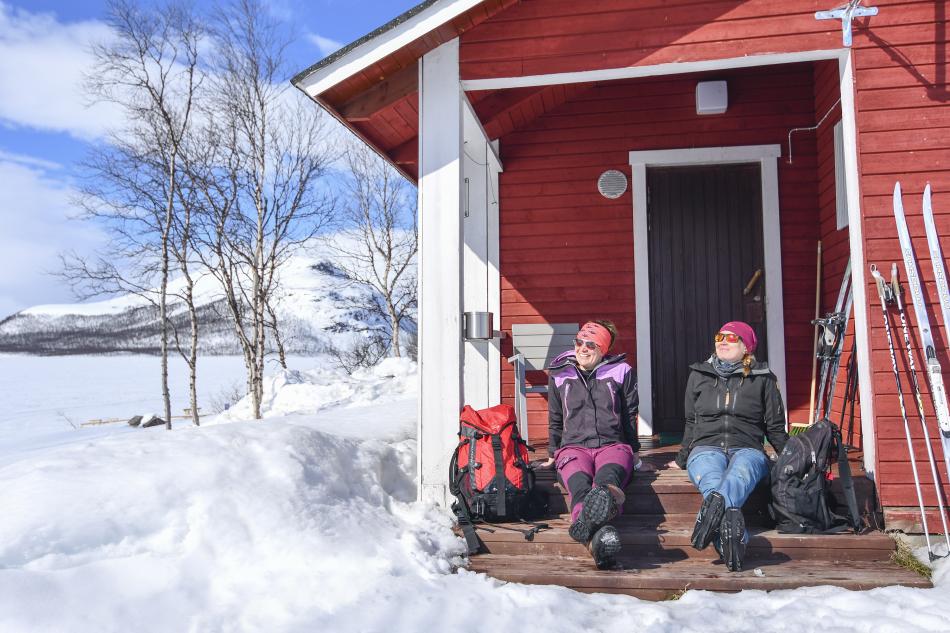 Punainen mökki Kilpisjärven rannalla keväisessä auringonpaisteessa. Taivas on sininen ja kaksi naista ottaa aurinkoa istuen mökin portailla. Tunturisukset nojaavat punaisen mökin seinään. 