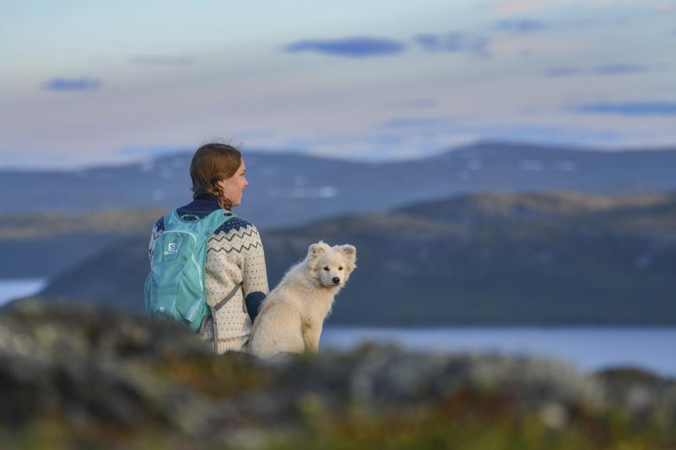 Nainen ja koira istuvat tunturin päällä mättäällä. Valkoinen koira katsoo suoraan kameraan ja nainen reppu selässään ihastelee maisemia.